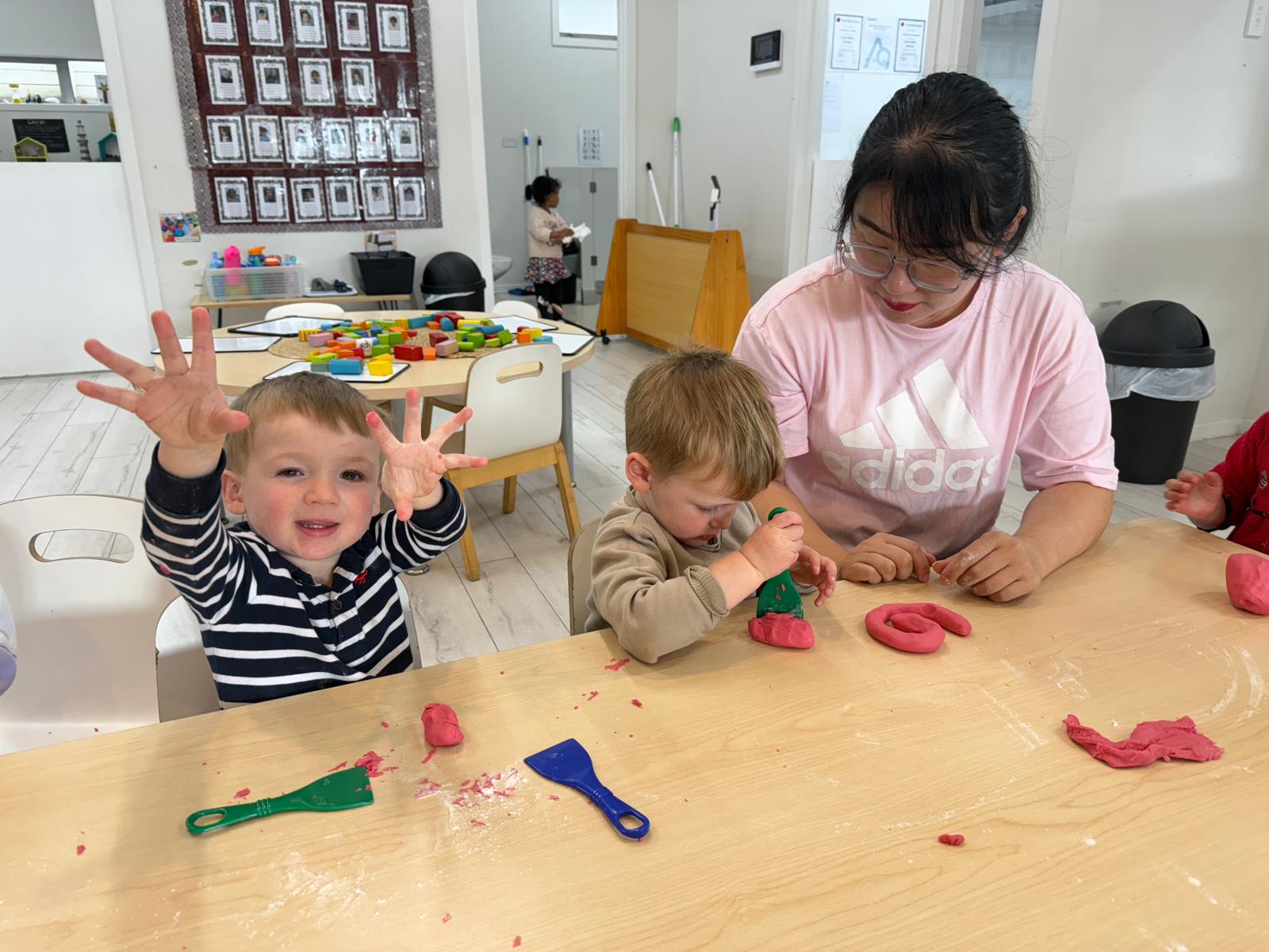 Toddler Room Team Leader Lu Li leads a creative playdough session with two toddlers at Bright Beginnings Mount Roskill, as one child smiles and waves playfully at the camera